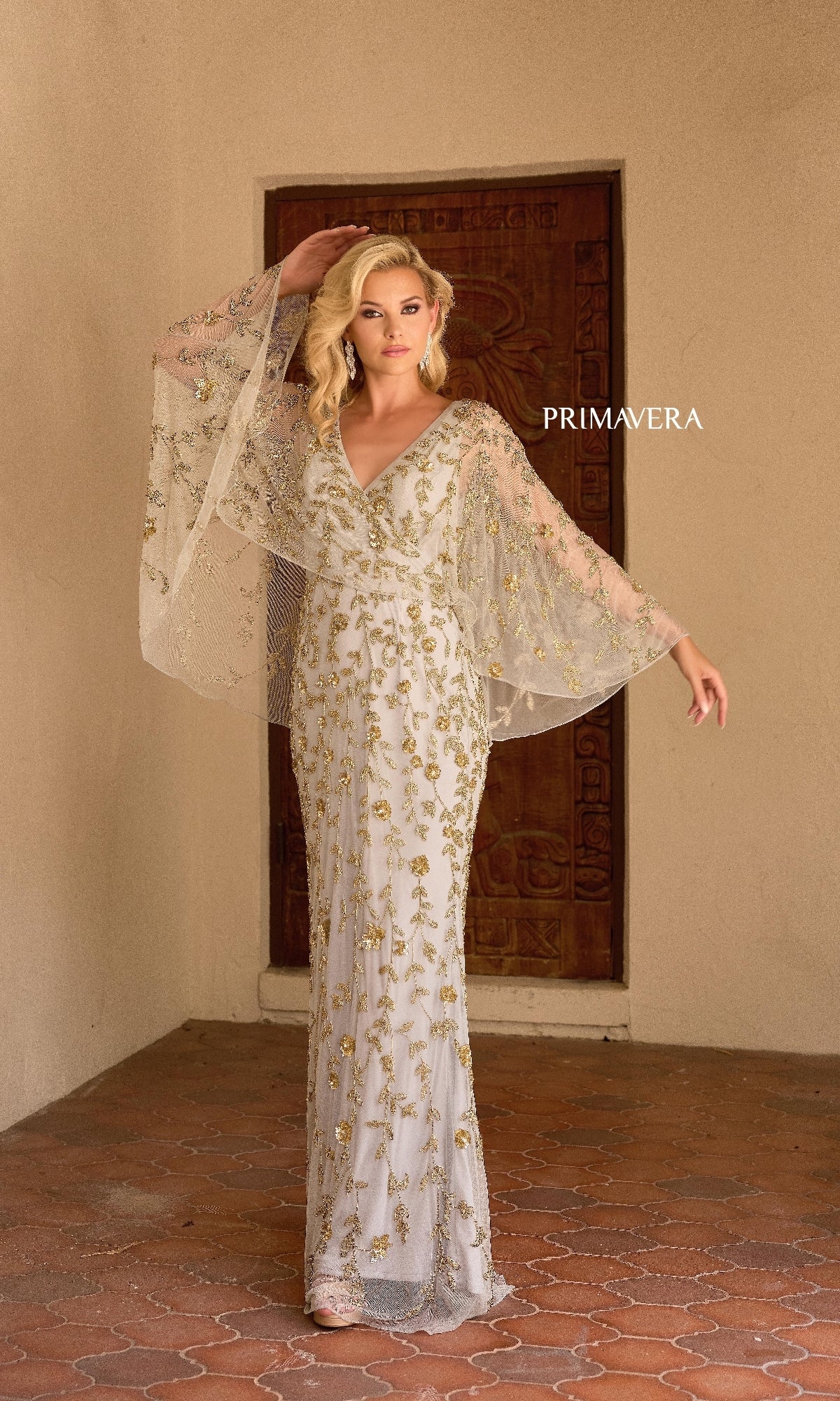 A woman stands before a wooden door wearing the Primavera 13149 long formal dress, featuring gold beading, sheer wide sleeves, and a v-neckline. The word 