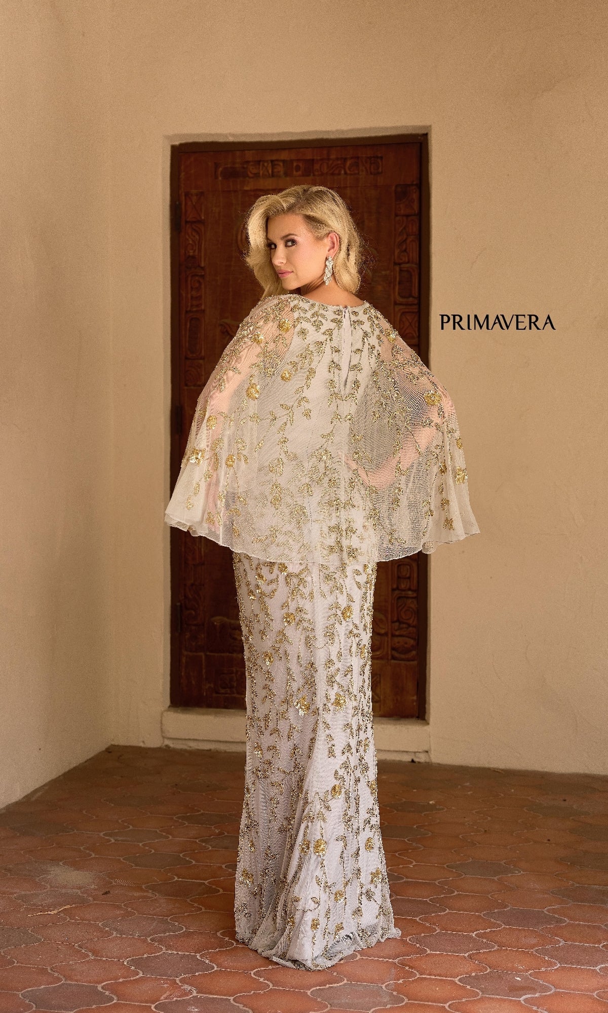 A woman wearing the Primavera 13149 long formal dress in white and gold beadwork with a capelet stands by a wooden door, looking over her shoulder, with 