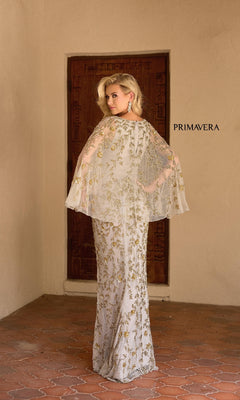 A woman wearing the Primavera 13149 long formal dress in white and gold beadwork with a capelet stands by a wooden door, looking over her shoulder, with "PRIMAVERA" on the wall behind her.
