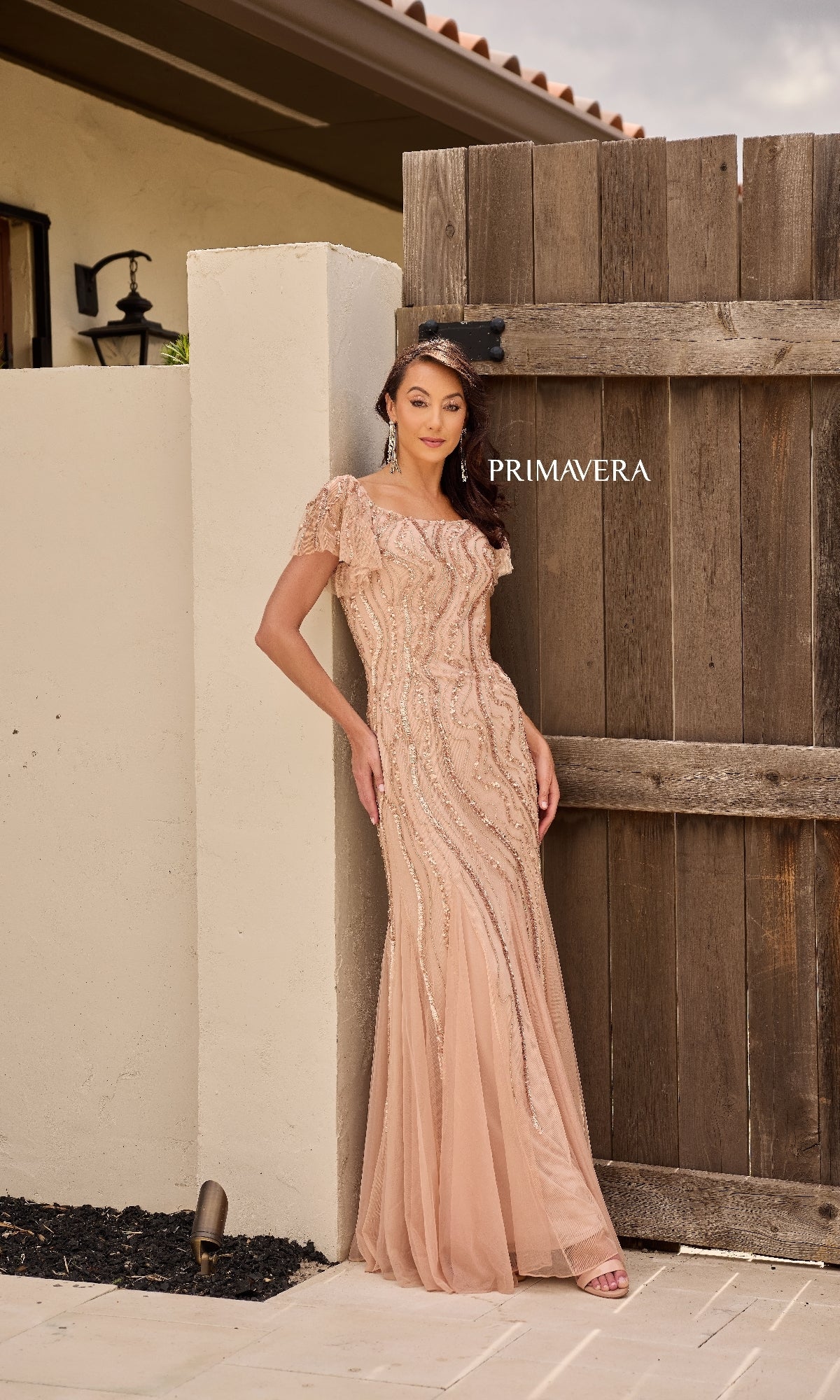 A woman poses outdoors against a wooden fence in the Primavera 13155 long formal dress, featuring an embellished off-shoulder blush design. The brand name 