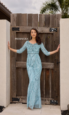 A woman wearing the Primavera 13156 long formal dress in light blue, featuring a beaded waistline, stands with arms outstretched, leaning on a wooden gate.