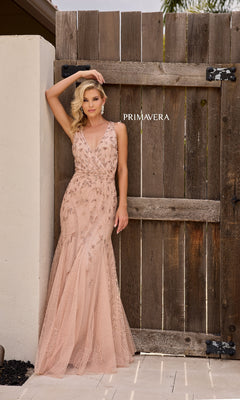 A woman models the Primavera 13158 long formal dress—a blush pink, embellished fit-and-flare gown with a v-neckline—posing outdoors against a wooden gate with "PRIMAVERA" text in the background.