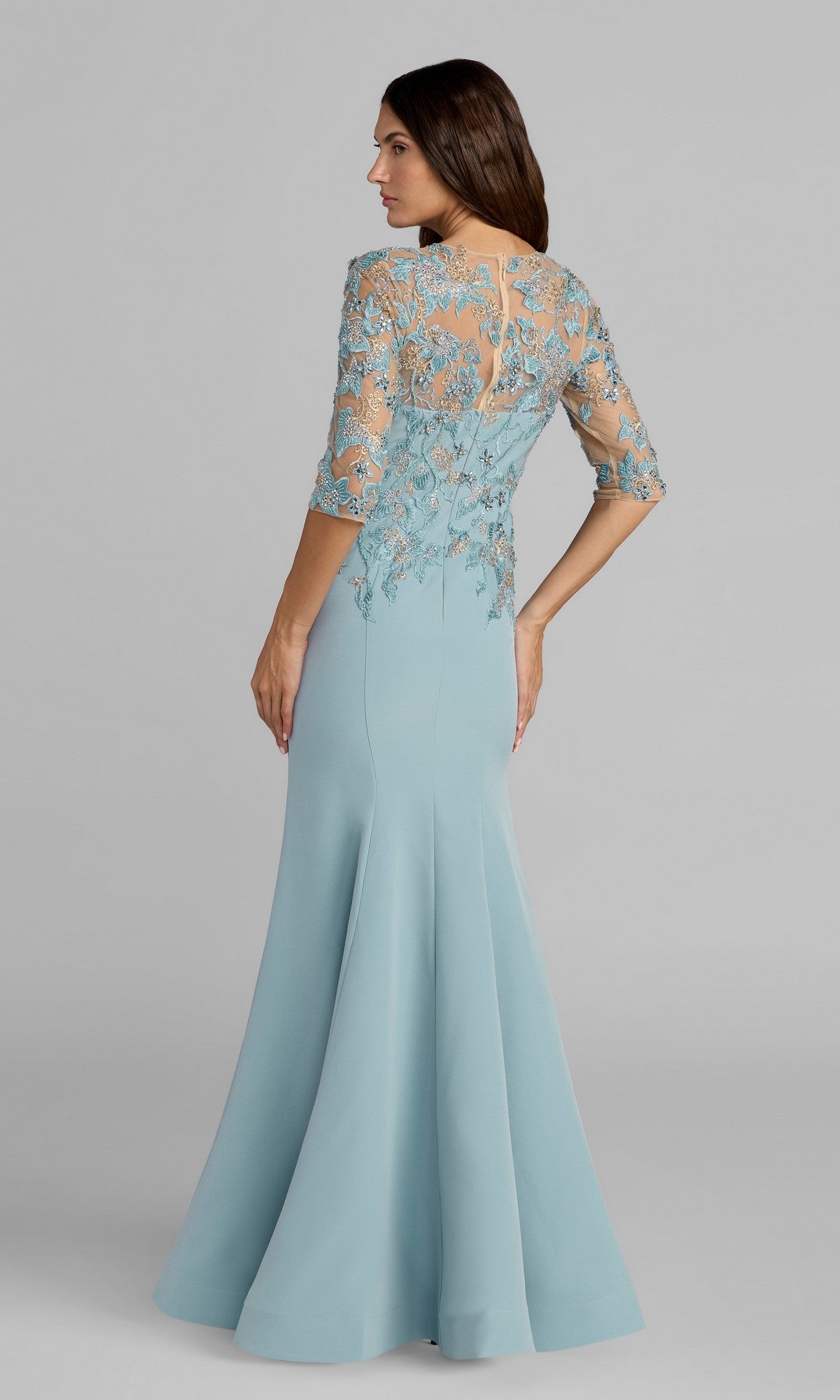 A woman stands with her back to the camera, wearing the Mac Duggal 13204 long formal dress in light blue, featuring lace and floral embroidery on the upper half and a floor-length silhouette.
