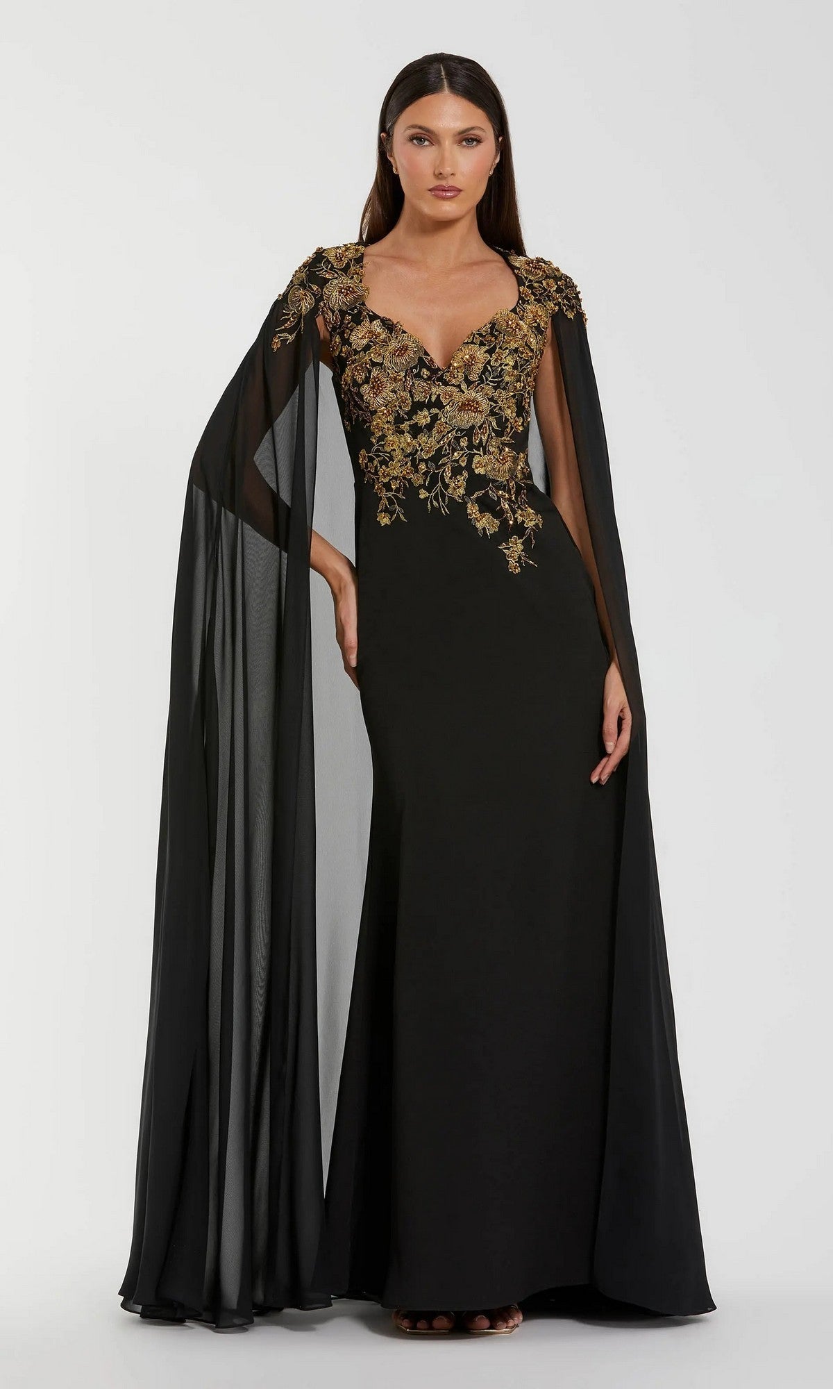 A woman wearing the Mac Duggal 13206 long formal dress in black gold, featuring a floor-length gown with sheer cape sleeves and intricate gold embroidery on the bodice, stands against a plain light background.