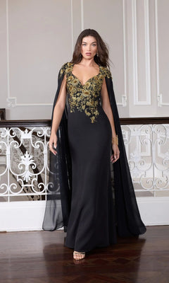 A woman wearing the Long Formal Dress: Mac Duggal 13206, featuring a long black gown with gold embroidery and a sheer black cape, stands indoors on a dark wooden floor beside an ornate white railing.