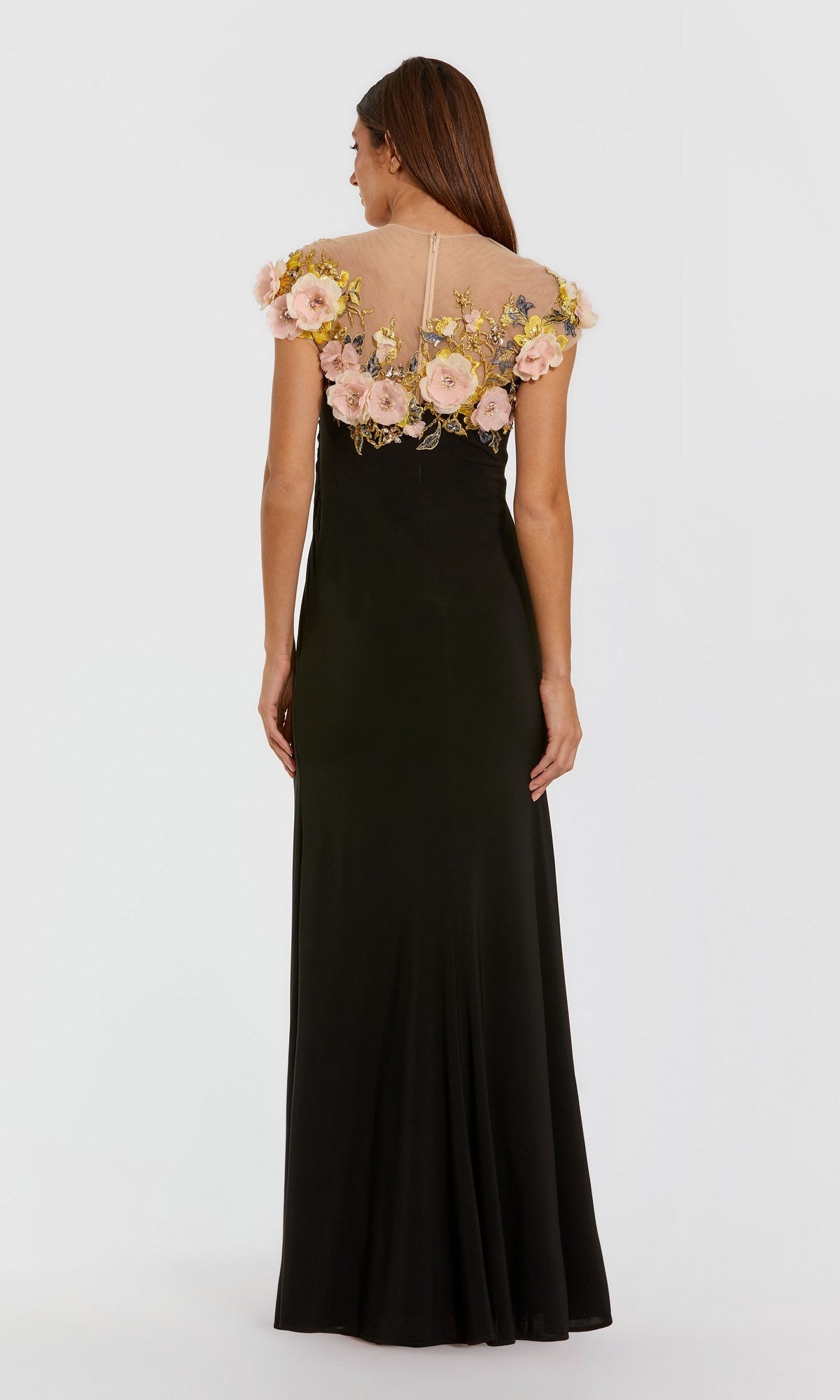 A woman stands facing away, wearing the Mac Duggal 13207 long formal dress—a black gown featuring short sleeves and delicate pink 3D floral accents that adorn the sheer upper back and shoulders.