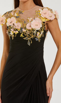 A woman is wearing the Mac Duggal 13207 long formal dress, featuring a black sheer neckline, pink 3D floral accents, gold embroidery, and cap sleeves. Only her upper body is visible.