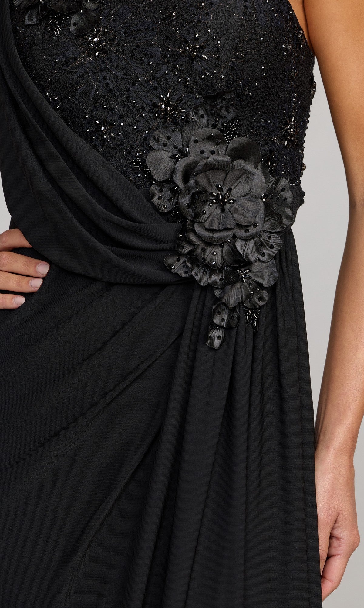 A person wearing the Mac Duggal 13210 long formal dress in black, featuring a one-shoulder design with floral appliqué, intricate beaded embellishments, and striking 3D flower detailing, shown from the shoulder to the hip.