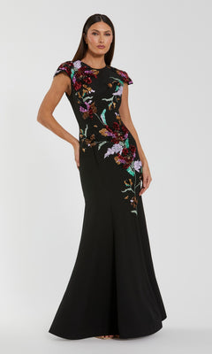 A woman stands against a plain light gray background wearing the Mac Duggal 13211 Long Formal Dress in Black Multi, featuring a fitted black crepe trumpet silhouette with vibrant floral sequin embroidery.