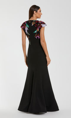 A woman stands facing away from the camera, wearing the Mac Duggal 13211 long formal dress in black crepe, featuring colorful floral sequin embroidery on the shoulders and upper back.