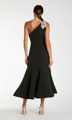A woman stands with her back to the camera, wearing the Tea-Length Party Dress: Mac Duggal 13221 in black crepe, featuring a one-shoulder design adorned with an embellished floral shoulder applique. She pairs the dress with beige high-heeled sandals.