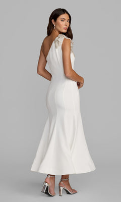 A woman in the Tea-Length Party Dress: Mac Duggal 13221, a fitted crepe one-shoulder midi dress with an embellished floral shoulder applique, paired with silver high-heeled sandals, stands against a plain gray background.