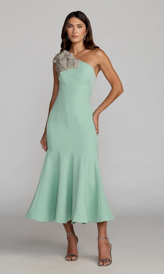 A woman wearing the Mac Duggal 13221 tea-length party dress in sage, a crepe one-shoulder design featuring a large embellished floral applique at the shoulder, stands against a plain gray background.