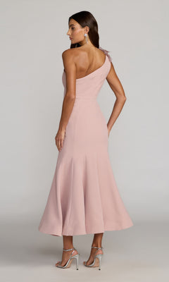 A woman stands facing away, wearing the Tea-Length Party Dress: Mac Duggal 13222 in blush pink crepe with a one-shoulder neckline, flared hem, and delicate 3D floral embellishments, elegantly paired with silver high-heeled sandals.