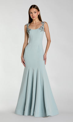 A woman stands against a plain light background, wearing the Mac Duggal 13224 Long Formal Dress in powder blue—a floor-length gown featuring floral embellishments on the straps.