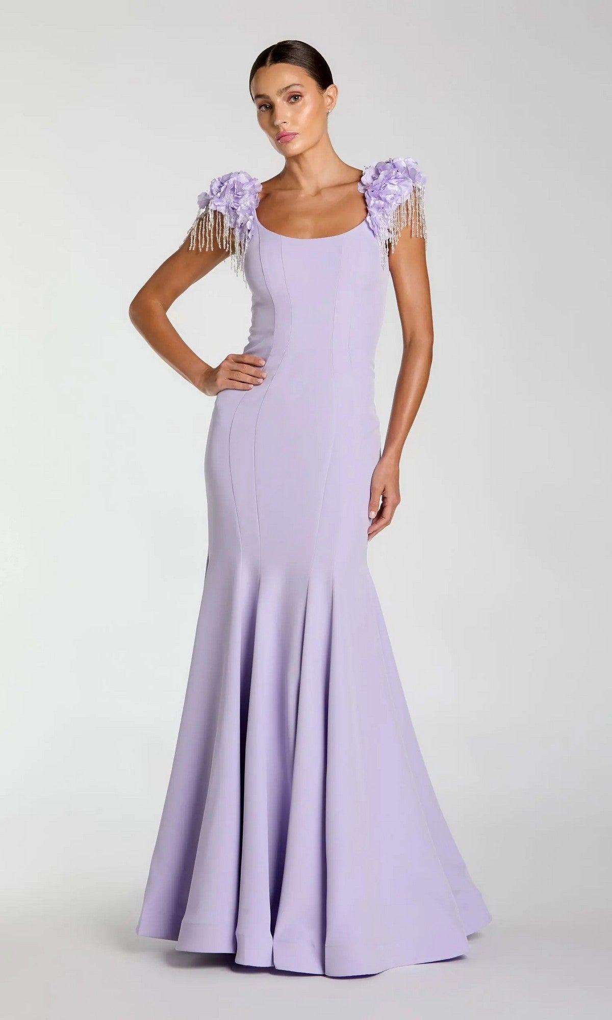 A woman models the Mac Duggal 13232 Long Formal Dress in lilac—a floor-length, fitted gown featuring floral and beaded shoulder embellishments and a graceful mermaid silhouette—posed against a plain light gray background.
