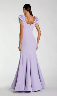 A woman faces away from the camera against a plain background, wearing the Mac Duggal 13232 long formal dress—a fitted, floor-length purple gown featuring a mermaid silhouette with a flared hem and floral shoulder embellishments.