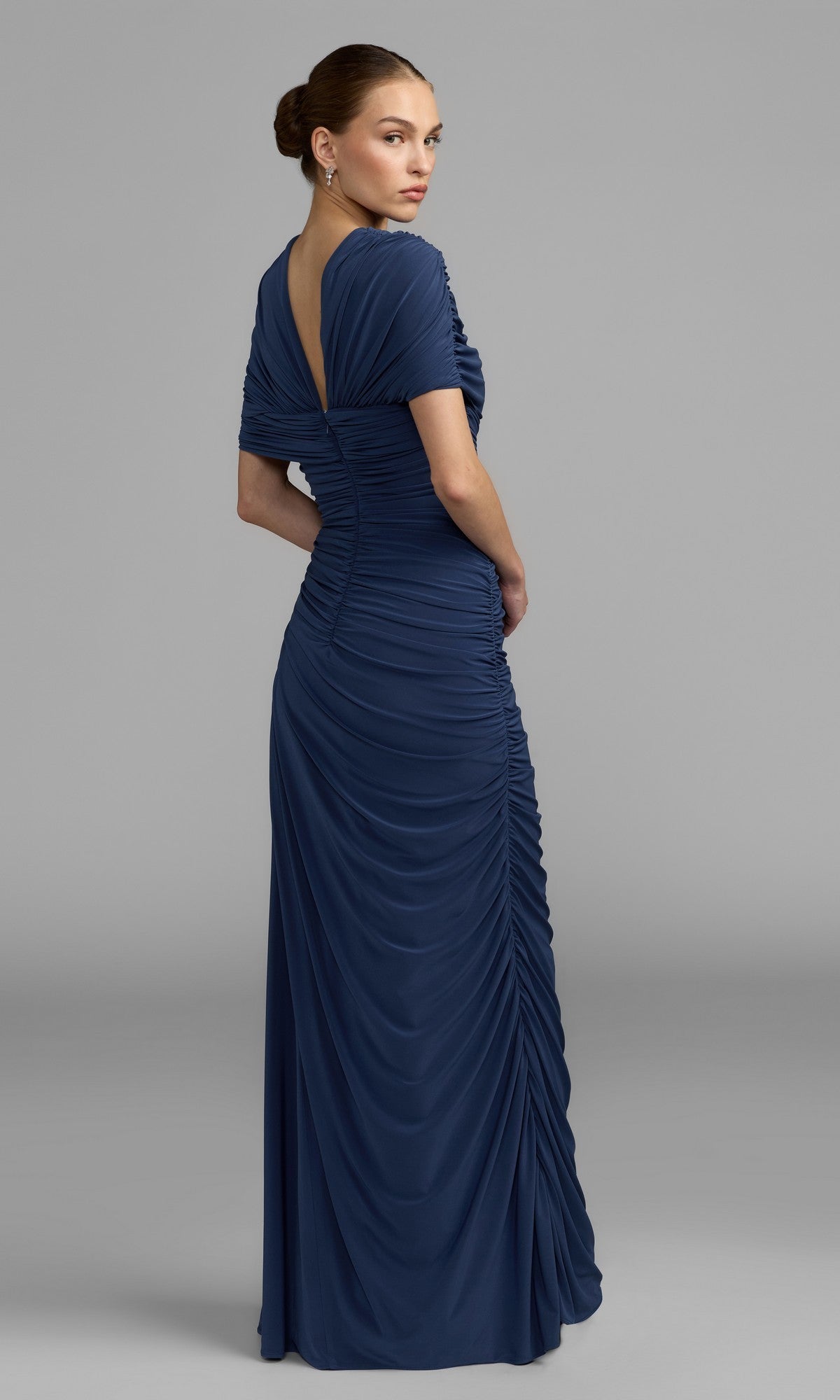 Woman in the Mac Duggal 13244 long formal dress—a floor-length, dark blue gown featuring ruching and short sleeves—standing sideways and looking over her shoulder against a plain gray background; an elegant and stunning option for a mother-of-the-bride seeking a sophisticated blue dress.