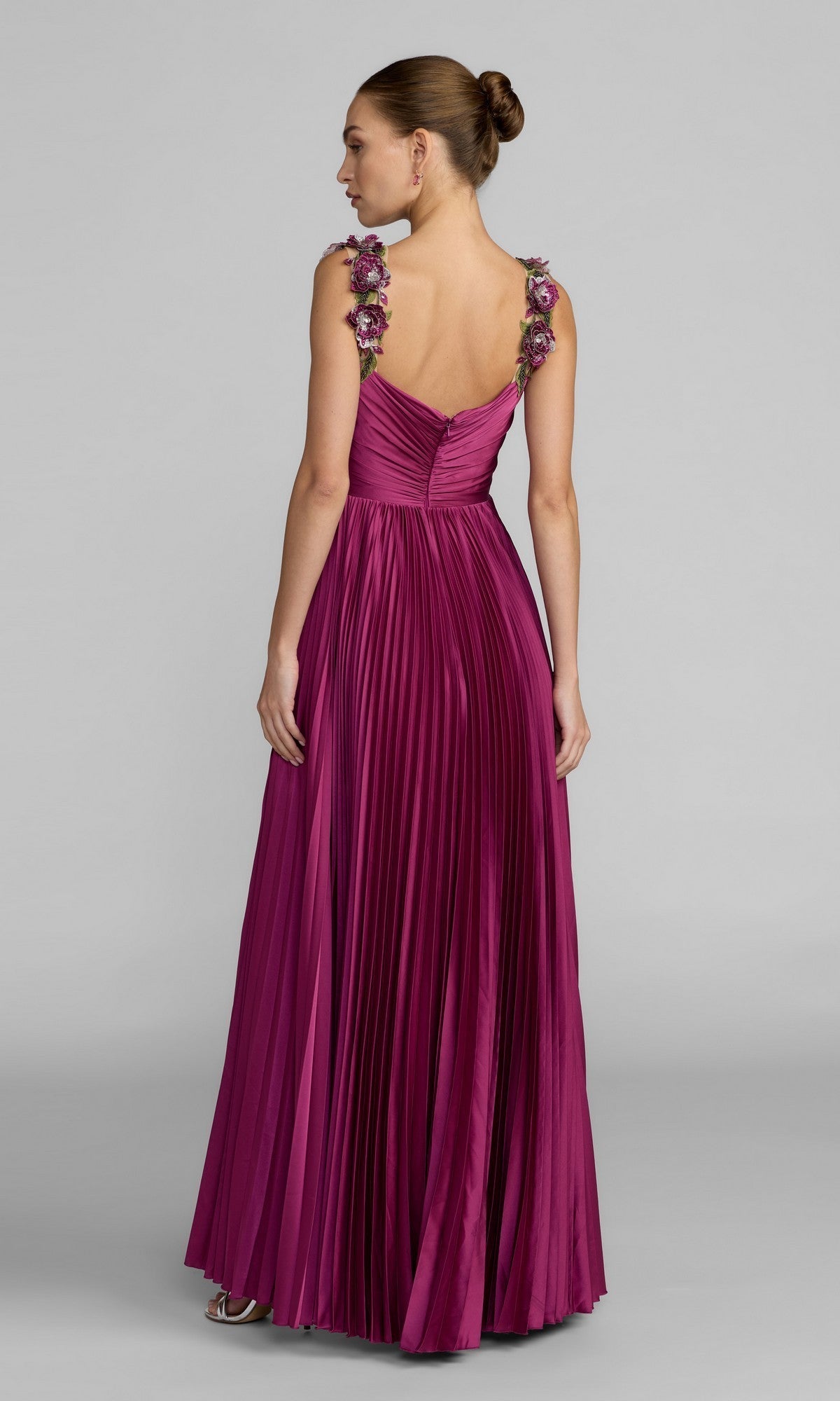 A woman stands with her back to the camera against a plain gray background, showcasing the Mac Duggal 13263 long formal dress in magenta satin, featuring a floor-length pleated skirt and delicate 3D floral straps.