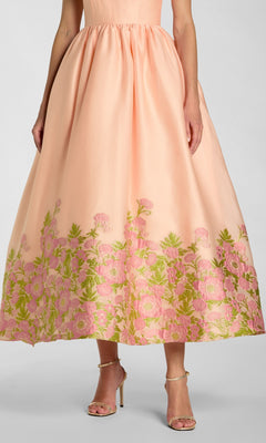 A woman wearing the Mac Duggal 13266 tea-length party dress in peach organza, featuring a sleeveless bodice and a full skirt with floral-embroidered hem in shades of pink and green; paired with strappy heels.