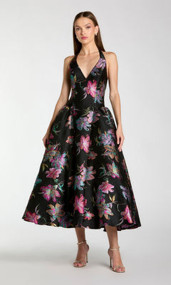 A woman wearing the Mac Duggal 13297 Tea-Length Party Dress in Black Multi—a sleeveless brocade dress featuring a colorful floral pattern, V-neckline, and midi-length skirt—paired with silver high-heeled sandals.