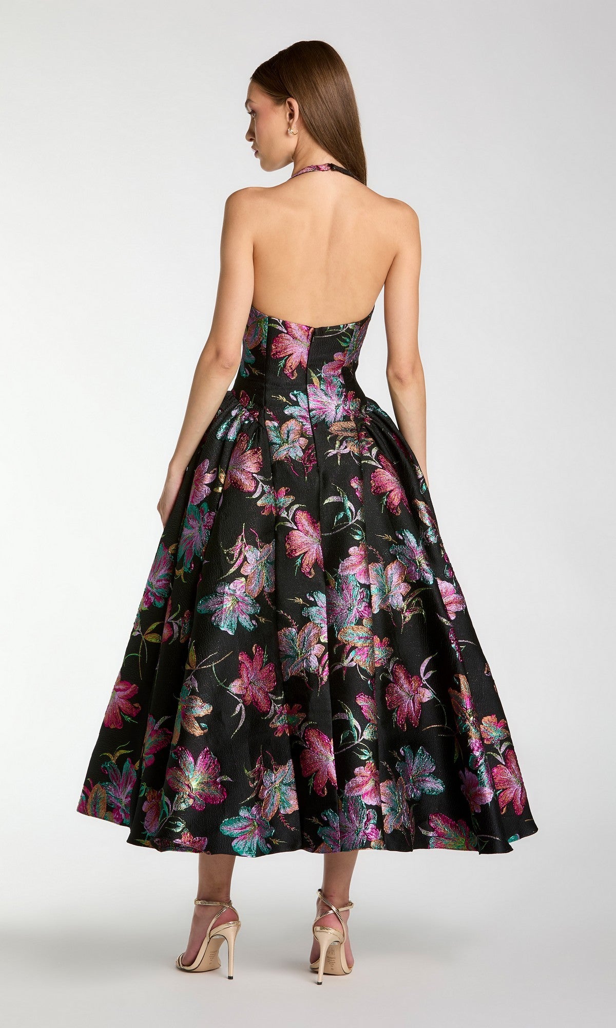A woman stands with her back to the camera, wearing the Mac Duggal 13297 Tea-Length Party Dress—an A-line, black halter-neck design featuring a floral print—paired with beige high-heeled sandals.