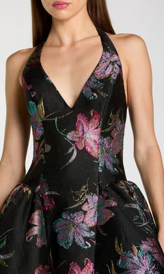 A woman models the Mac Duggal 13297 tea-length party dress, featuring a sleeveless design and deep V-neck, crafted from black floral brocade with a colorful pattern, as she poses against a plain background.
