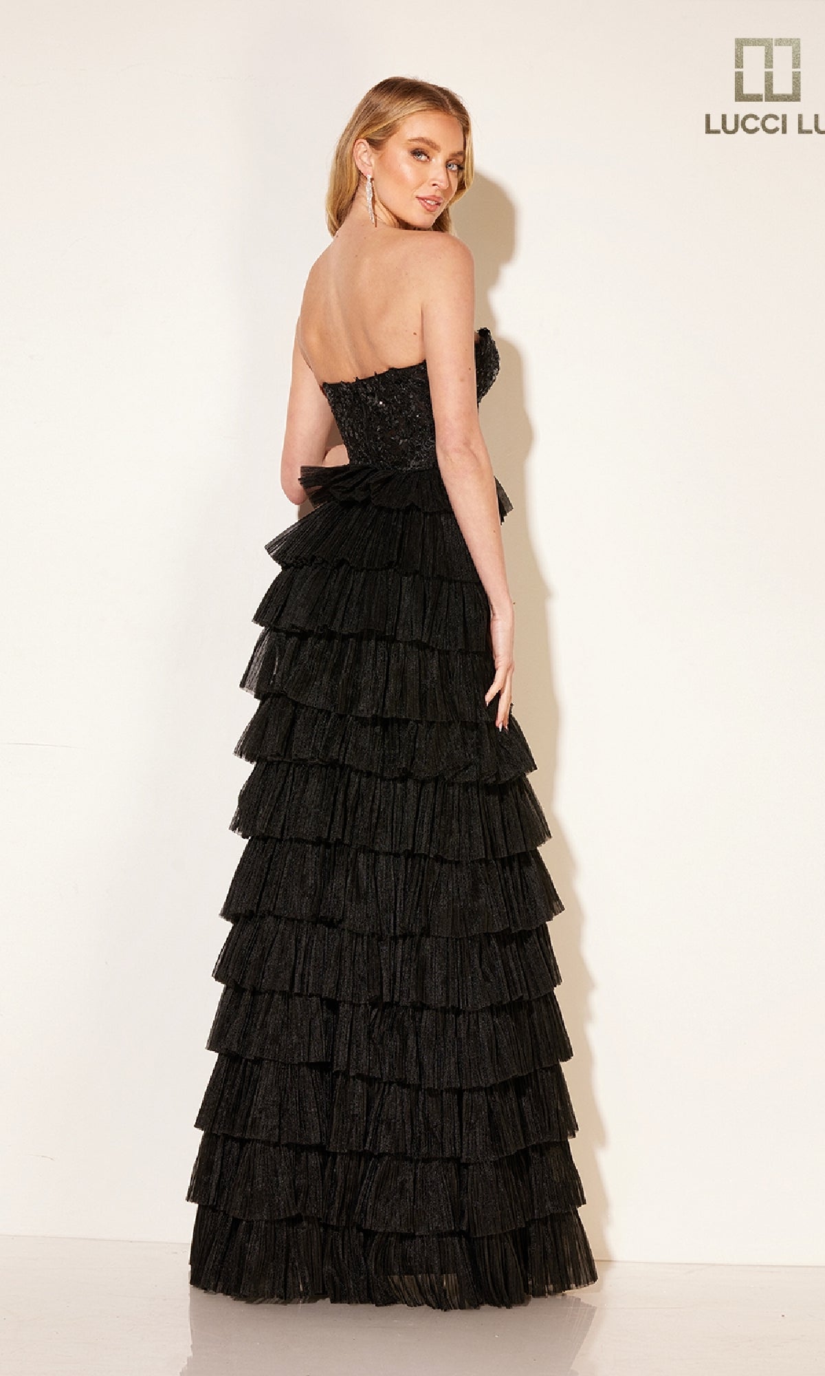 A woman wears the Lucci Lu 1344 Tiered A-Line Ruffle Prom Dress, a black strapless gown with multiple horizontal ruffled layers. She stands facing away, glancing over her shoulder against a plain white background.