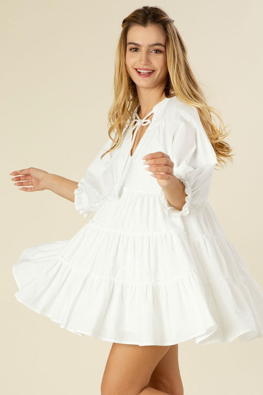A woman smiles and spins in a Short Off-White Casual Summer Dress with 3/4 sleeves and tiered design, set against a plain beige background.