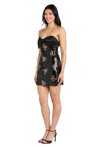 Woman wearing a black strapless short dress with pink bow sequin embellishments on a white background