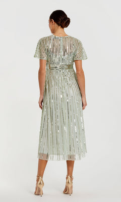 A woman stands facing away in the Mac Duggal 14007, a short sequin wedding-guest dress in light green with vertical metallic stripes and high heels—ideal for semi-formal occasions.