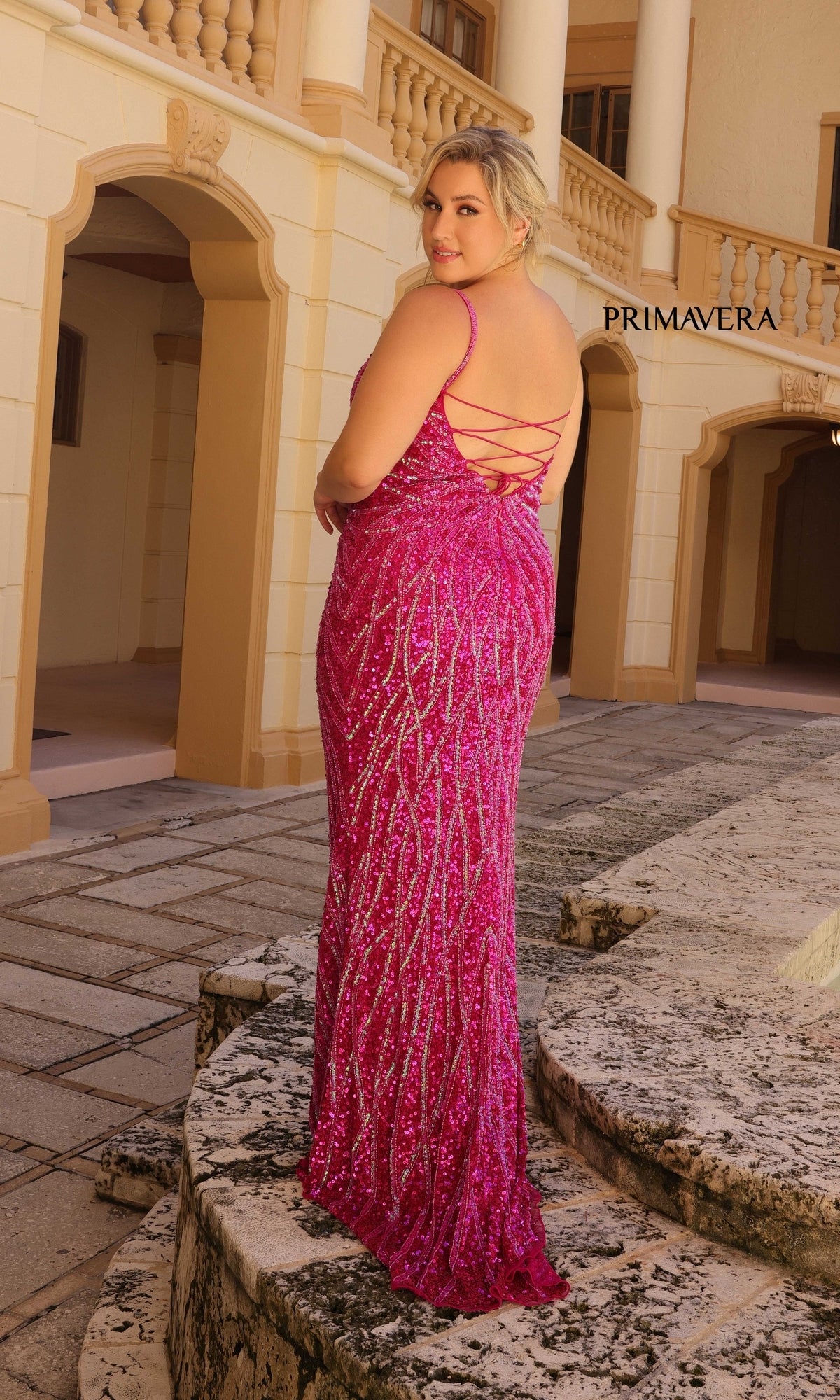 A woman wears the Primavera Plus-Size Long Beaded Prom Dress 14045, featuring bright pink sequins and a lace-up back, standing outdoors on a stone walkway by a building with columns and arches.