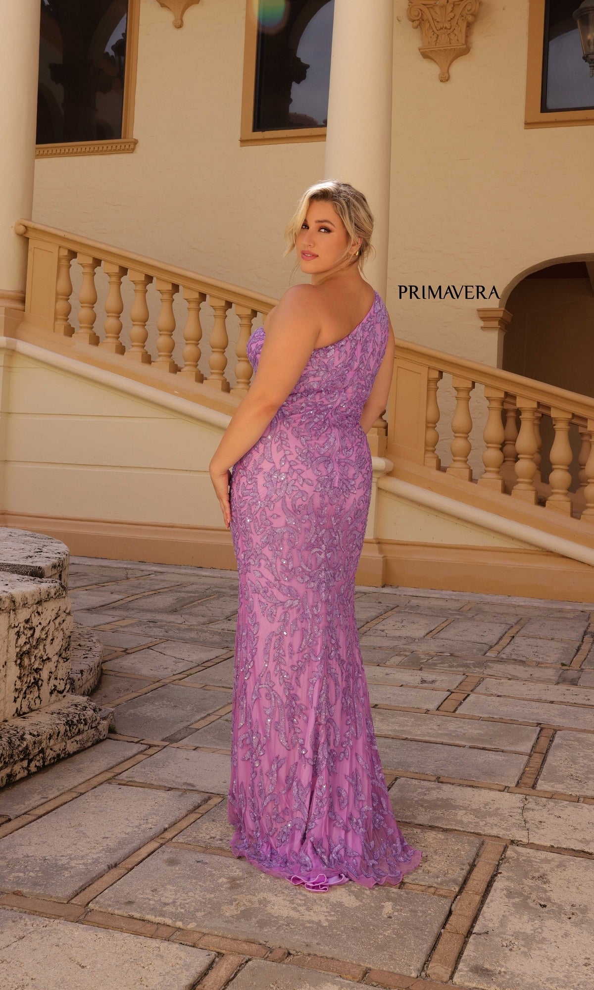 A woman in the Primavera Plus-Size Long Beaded Prom Dress 14049 stands on a stone patio in front of a building with columns and ornate railings.