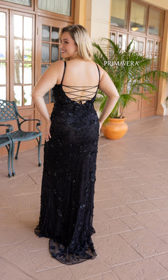 A woman wears the Plus-Size Long Beaded Primavera Prom Dress 14050, featuring black sequins and a lace-up open back, as she stands on a tiled patio by a potted plant and glass doors.