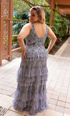 A woman stands on a tiled walkway in the Primavera Tiered Plus-Size Long Prom Dress 14052, with tiered ruffles and floral embroidery, facing away from the camera. Lush greenery appears in the background.