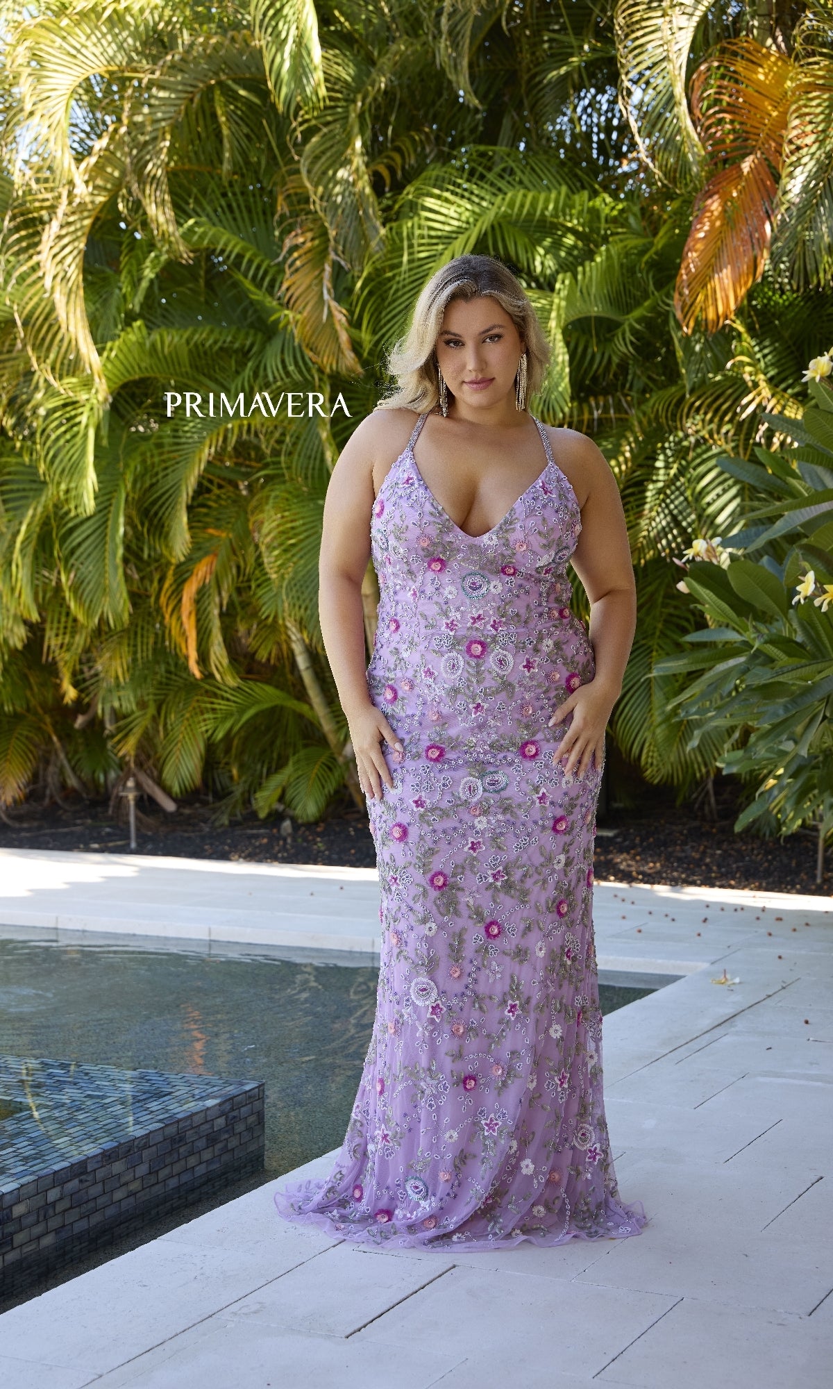 A woman wears the Plus-Size Beaded Prom Gown: Primavera 14071 in lavender floral, standing on a white patio by a pool with lush greenery behind her and 