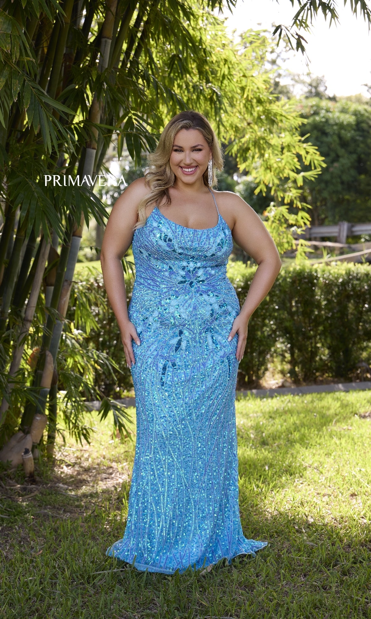 A woman stands on grass amid bamboo and lush greenery, wearing the Plus-Size Long Beaded Prom Dress: Primavera 14072—a sparkly light blue evening gown.