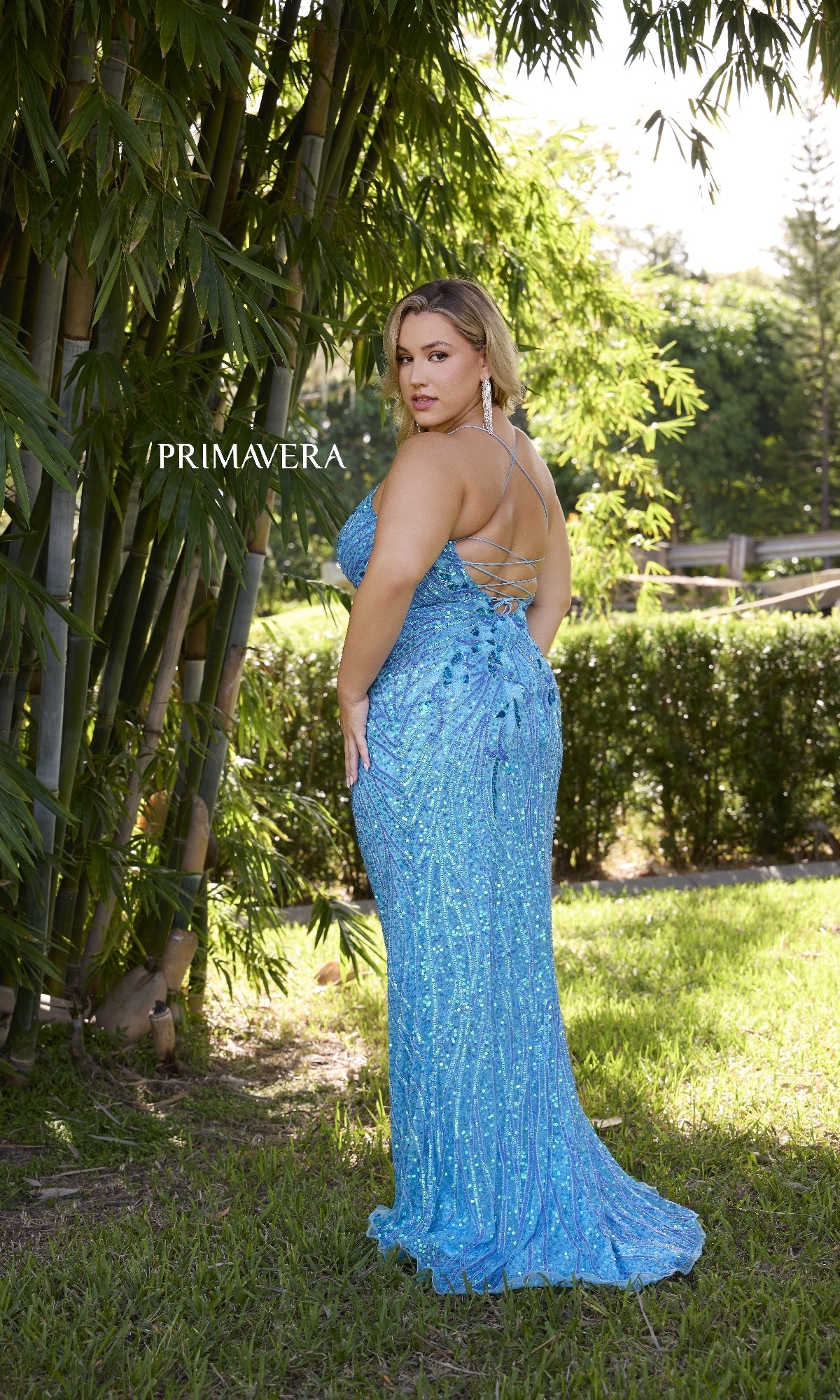 A woman in the Primavera 14072 plus-size long beaded prom dress stands on grass near bamboo, facing slightly away. The word 