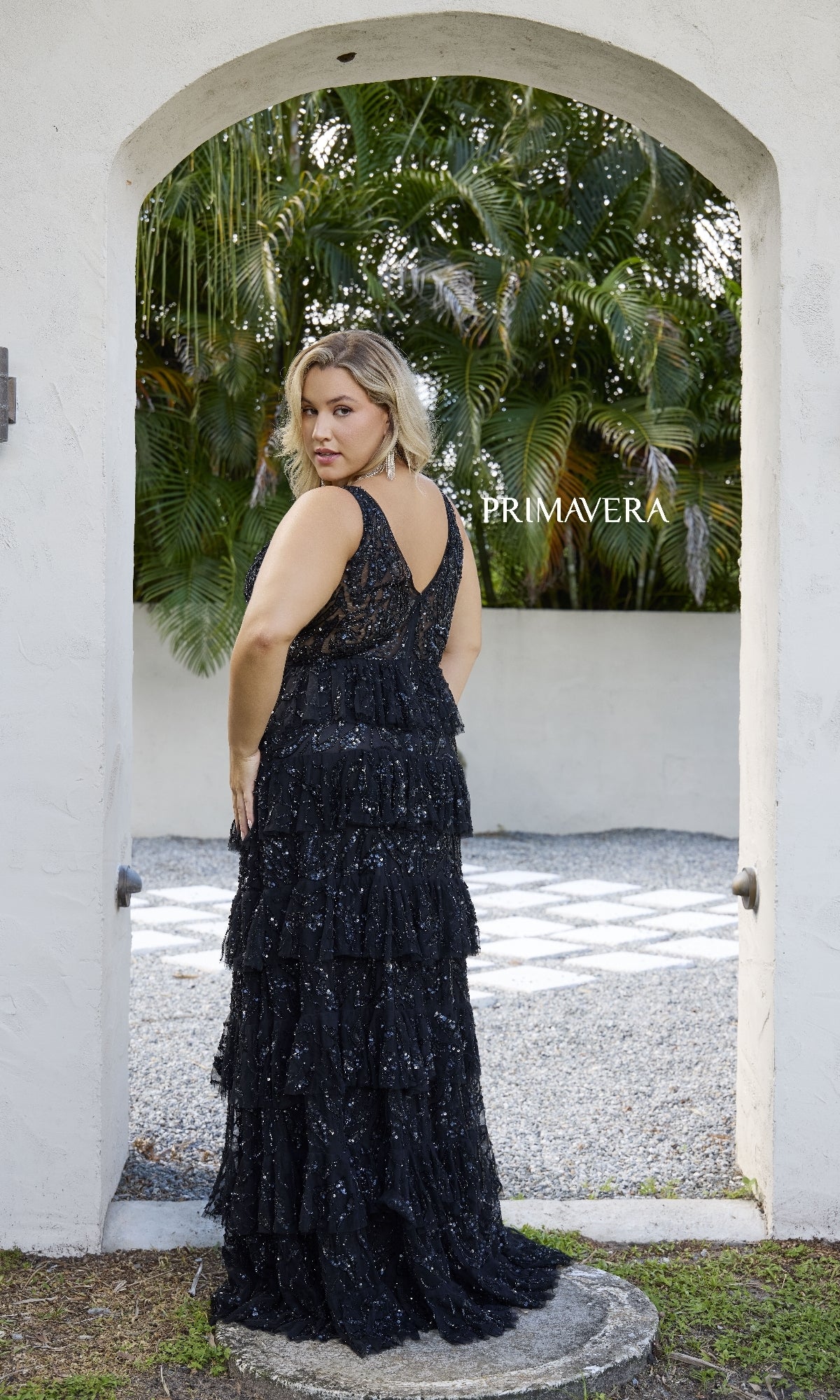 A woman models the Primavera 14073, a long plus-size black prom dress with sequin embellishments, standing outdoors under an archway and looking over her shoulder. The word 