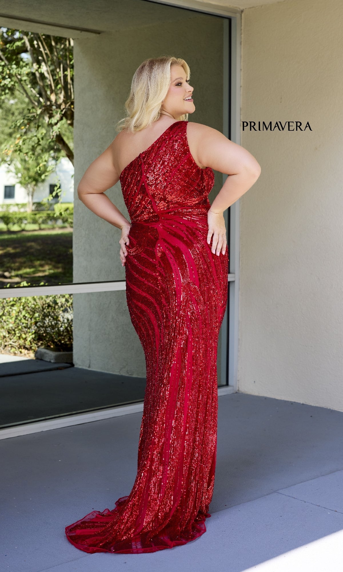 A woman wearing the Primavera 14075 Plus-Size Long Sequin Prom Dress in red stands on a patio near a glass door, facing away from the camera with 