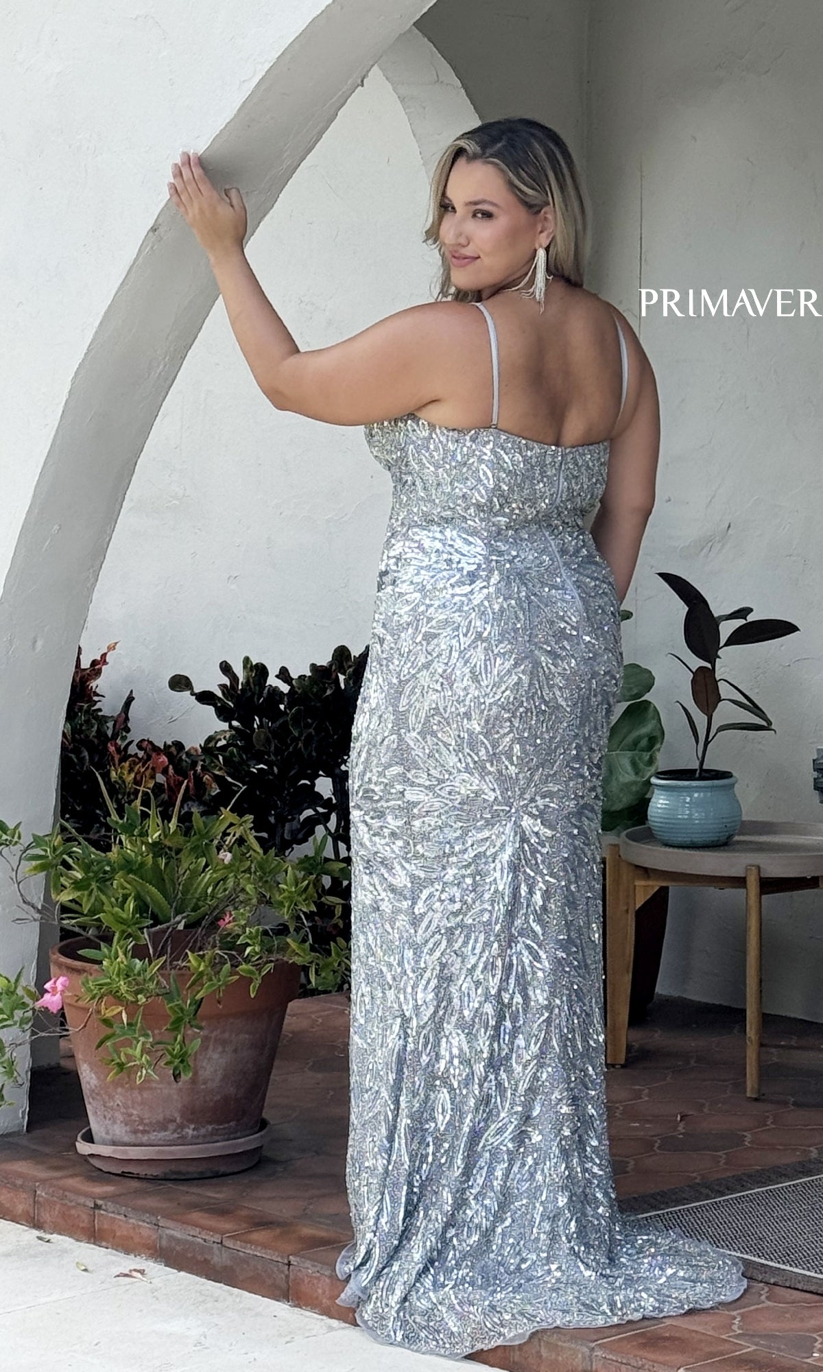 A woman wearing the Primavera 14078 plus-size long beaded prom dress stands on a porch near potted plants, facing away and glancing over her shoulder. 