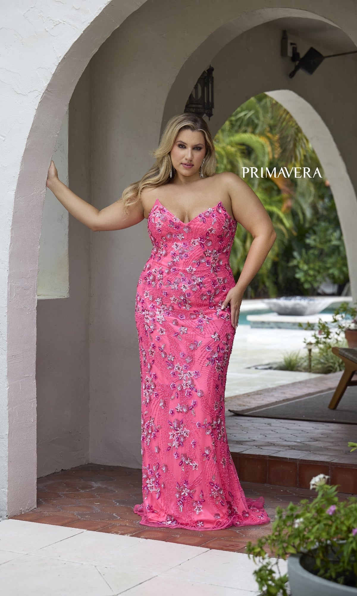 A woman in the Primavera 14080 Long Plus-Size Prom Dress stands under an archway with one arm resting on the wall, with 