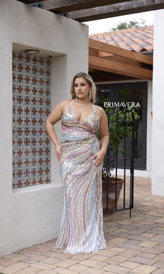 A woman in the Primavera 14082 Long Plus-Size Prom Dress, a multicolored sequin gown with a form-fitting silhouette, stands outside near a white wall with patterned tiles and a glass door labeled "Primavera.