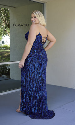 A woman stands outdoors near a window, wearing the Plus-Size Strapless Prom Gown: Primavera 14083—a stunning floor-length blue sequin dress with a lace-up back, perfect for any special occasion.
