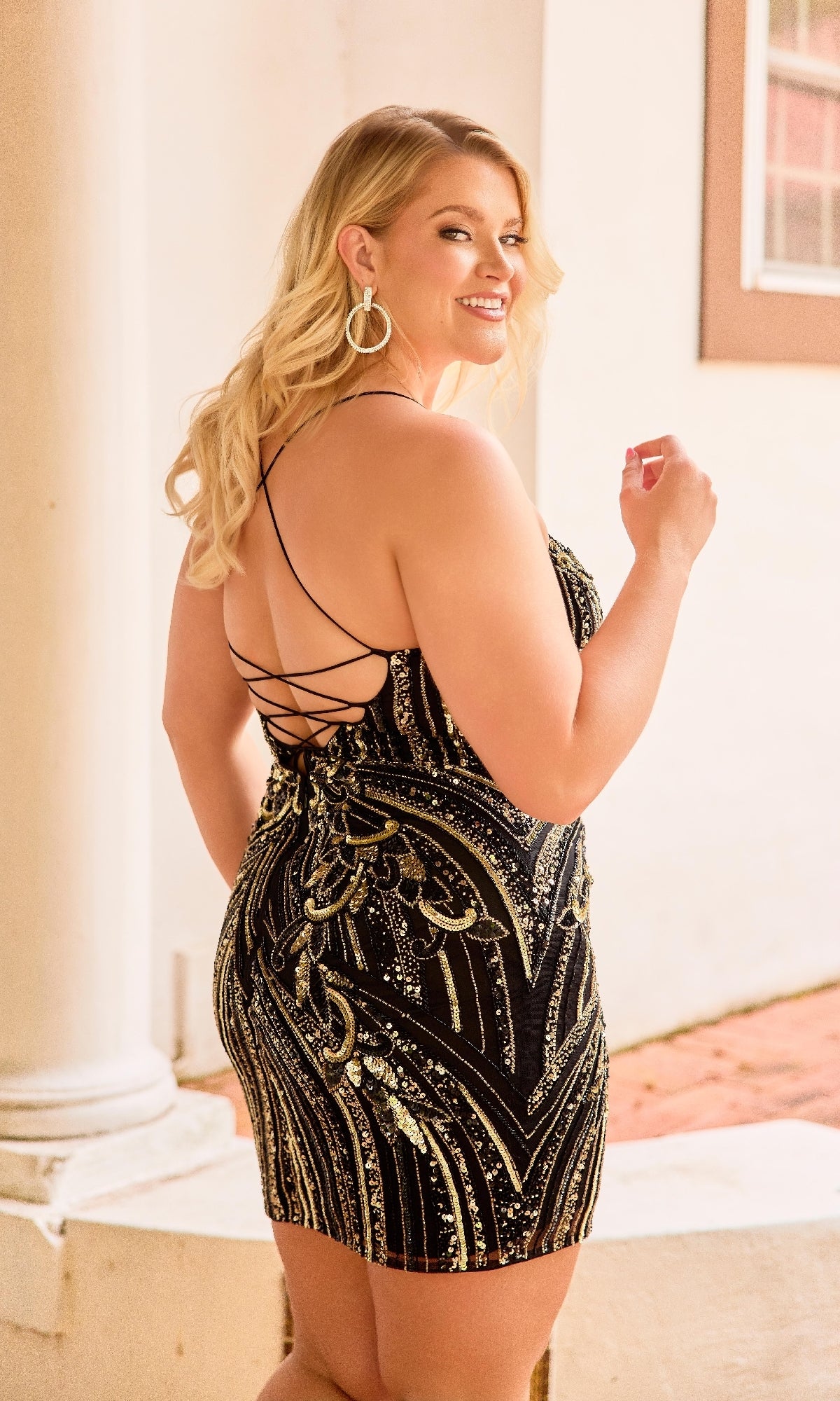 A woman with long blonde hair models the Plus-Size Mini Beaded Hoco Dress: Primavera 14093, featuring black and gold sequins and a strappy-back design, as she smiles over her shoulder by a white column.