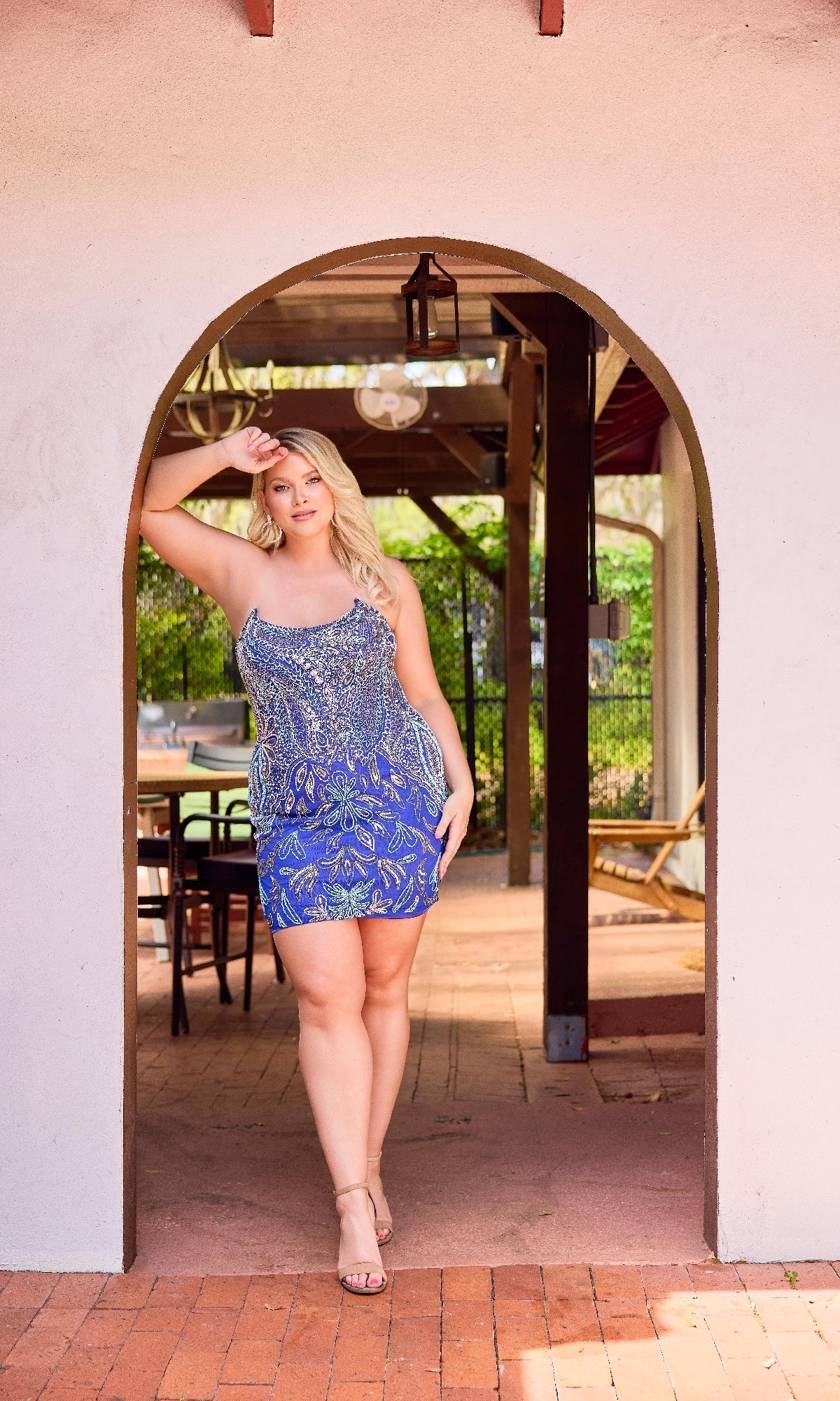 A woman wears the Primavera 14094 Plus-Size Strapless Hoco Dress and sandals, standing under a stucco archway with one hand on her forehead in an outdoor setting.