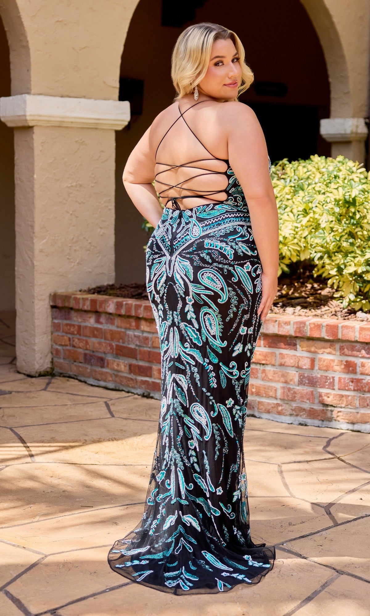 A woman stands outdoors in the Primavera 14102 long plus-size prom dress, featuring blue and silver beaded patterns and a fitted black floor-length design with an adjustable lace-up back.