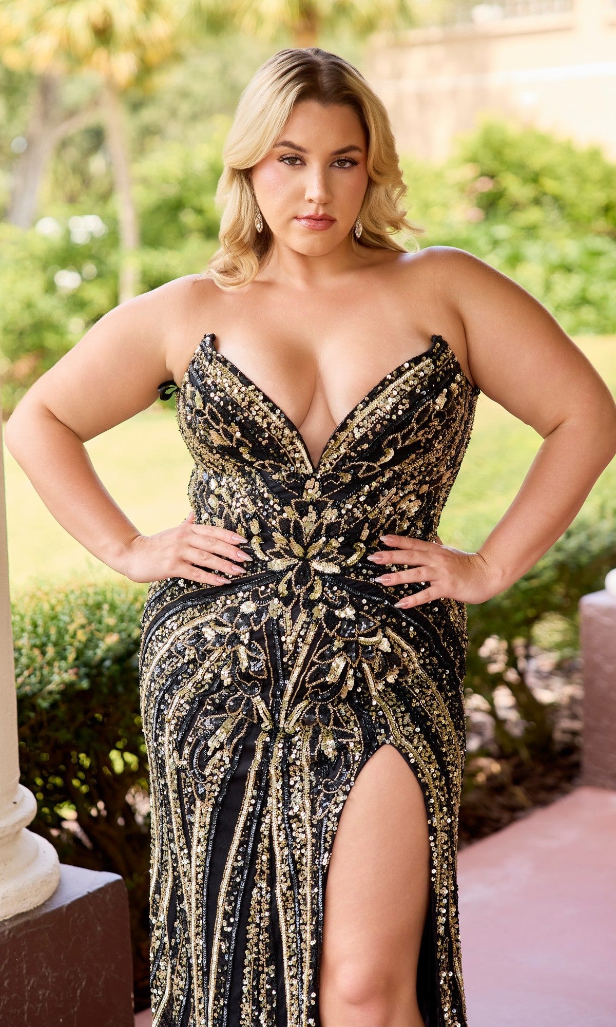A woman poses outdoors in the Primavera 14103 long plus-size prom dress—strapless, black and gold with shimmering sequins, beads, and a thigh-high slit. She stands confidently with hands on hips, wearing this glamorous formal evening gown.