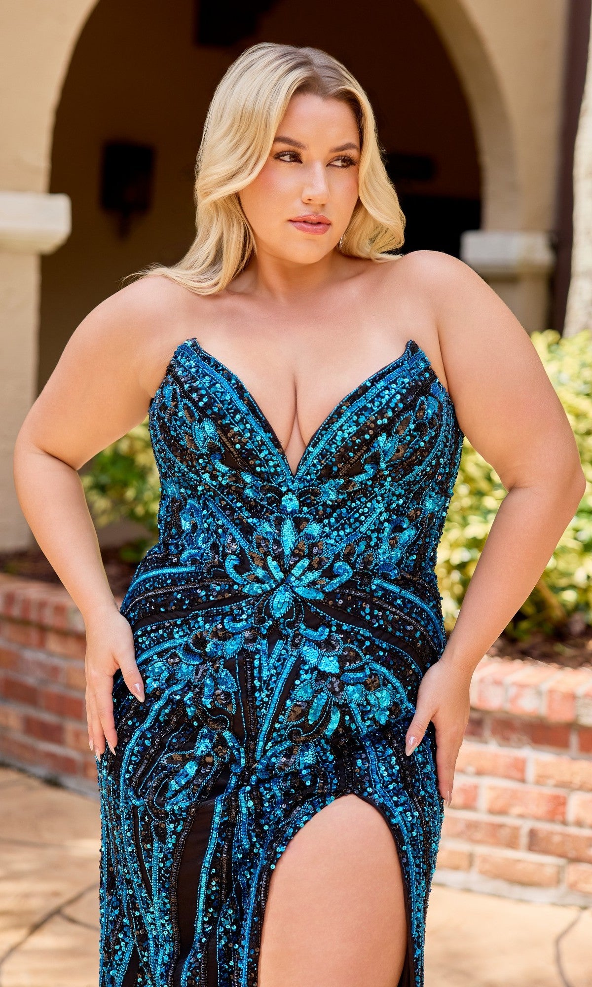 A woman stands outdoors in front of a brick and stucco building, wearing the Primavera 14103 long plus-size prom dress—a strapless black and blue sequin beaded gown with a thigh-high slit.