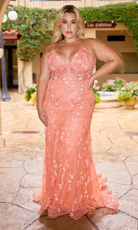 A woman stands on a stone patio in the Long Plus-Size Prom Dress: Primavera 14106, a floor-length coral gown with sparkly details and peach floral embroidery, featuring a deep V-neckline. Greenery and potted plants create a lush backdrop.